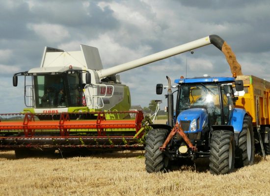 A modern combine harvester and tractor working together to harvest wheat in a sunny outdoor field.