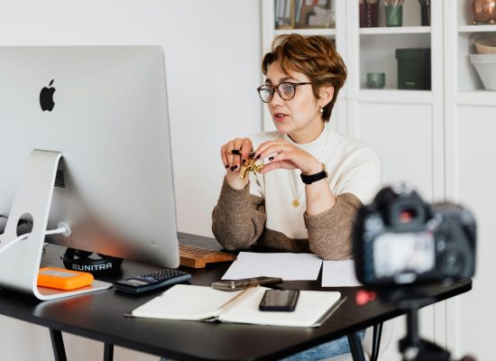 A professional woman engaged in a virtual meeting setup at her home desk, using a computer and camera.